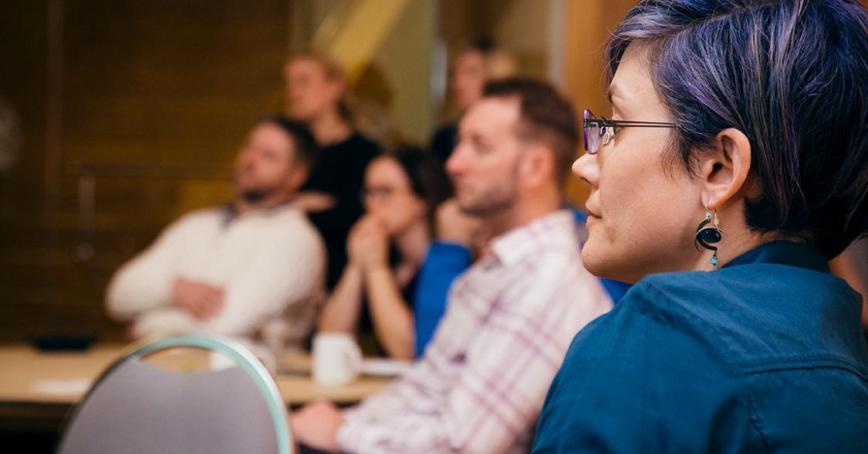 A group of people seated in a room, their attention is focused toward an (unseen) presenter. A woman with dark hair streaked with purple is in the foreground.