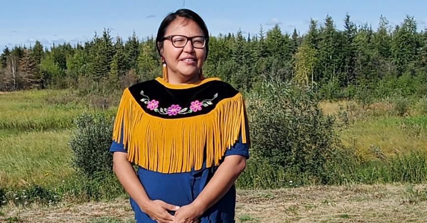 Chief-elect Doreen Arrowmaker stands outside with trees in the far background. She smiles softly and her hands are held at her waist. She is wearing a fringed leather shall with a band of black and pink flowers.