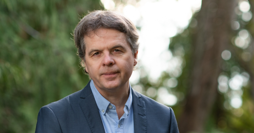 David Black poses with a neutral expression, wearing a blazer with a forested scene in the background.