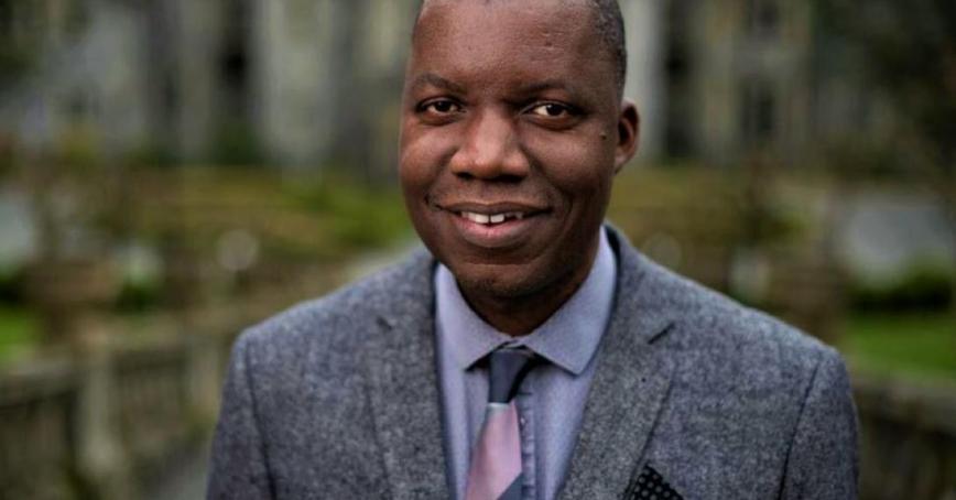 A man in suit and tie looks at the camera, offering a small smile. The background shows a stone building and greenery.