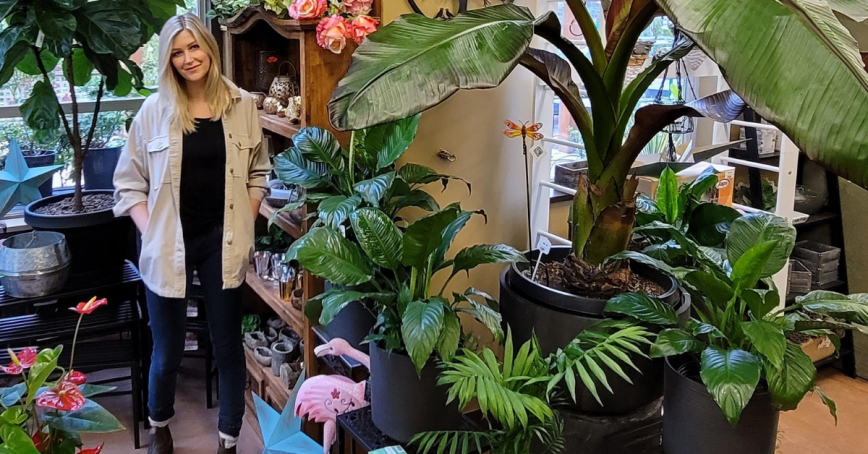 A woman stands in doors surrounded by towering potted plants and leafy greens.