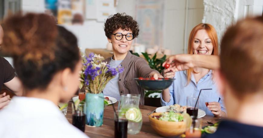 An image with several individual sitting at a table and one person is stretching a bowl to another person. 