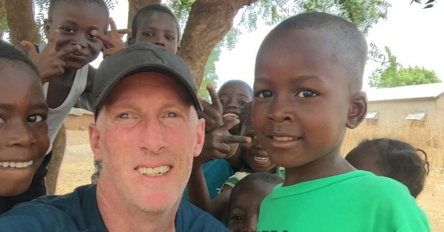 Man in baseball hat squats down so a group of seven children can also be seen, they are gathered around and smiling into the camera