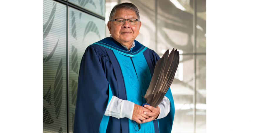 Lekwungen Elder Butch Dick standing in convocation regalia with traditional embroidery down the front, holding eagle feathers.