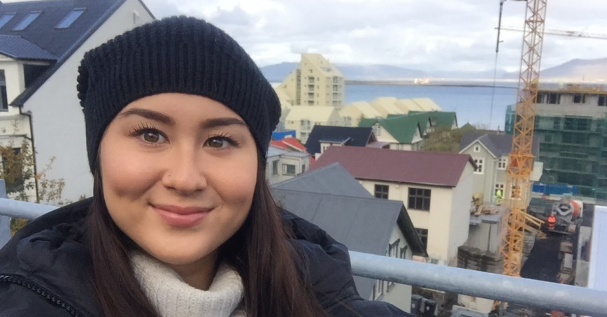 A woman in a toque smiles from what might be a balcony. Homes and a crane can be seen in the distance below.