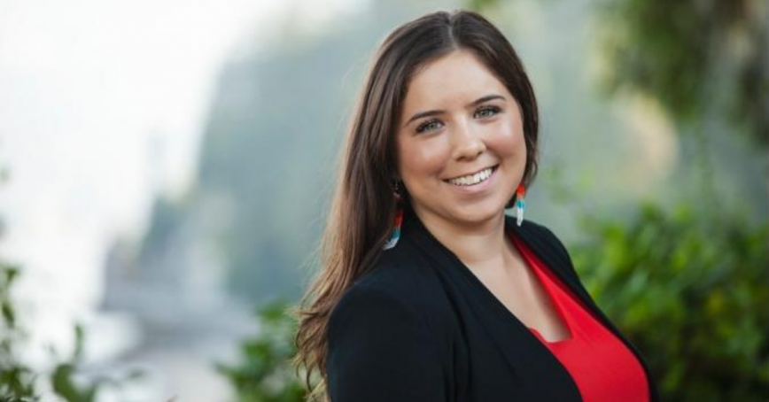 A smiling woman poses for the camera with a blurred out background of bushes and trees behind her.