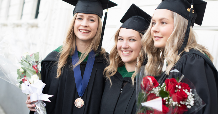 Three graduates smile while holding flowers in their caps and gowns. One is wearing a convocation medal.