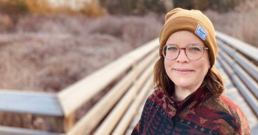 A woman in a toque smiles at the camera while standing on a boardwalk.