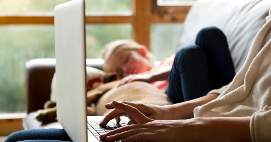 woman typing on laptop with child sleeping on couch beside her