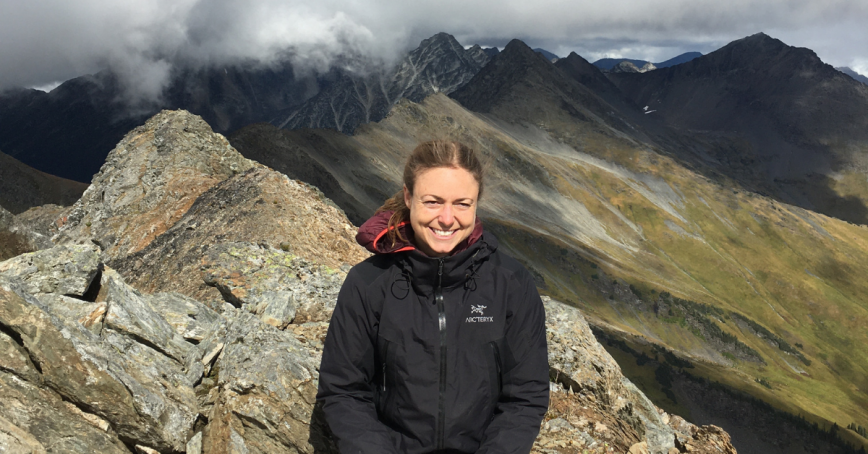 Kristina Schrange poses on a rocky peak wearing a black jacket. High altitude clouds and a green mountain slope are in the background.