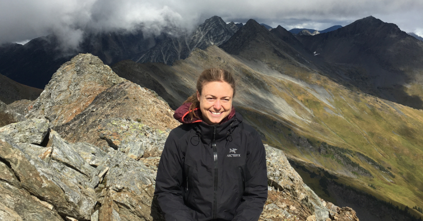 Kristina Schrange poses on a rocky peak wearing a black jacket. High altitude clouds and a green mountain slope are in the background. 