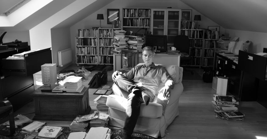 Black and white image. A man sits in a wingback chair surrounded by books. The sun streams in through the window.