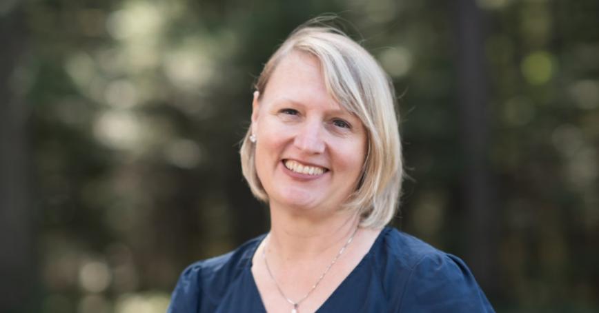 Tracy Smith-Carrier, a blonde woman with a big smile, stands outside with blurry green trees in the background.