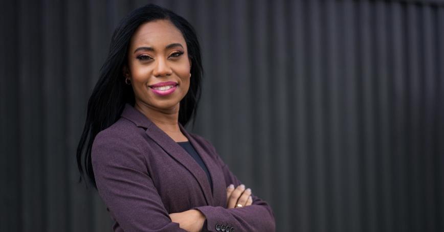 RRU grad Siobhan Calderbank, a Black woman in a purple suit jacket, crosses her arms and smiles. 