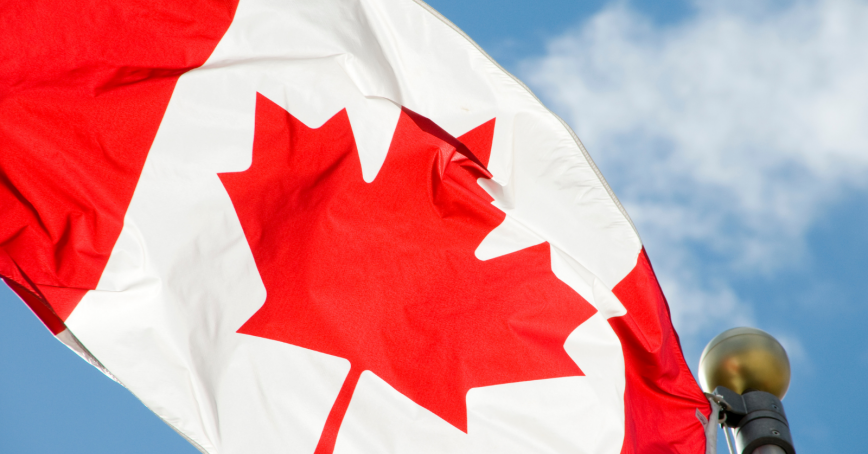 The Canadian flag waves in front of a blue sky with fluffy white clouds.