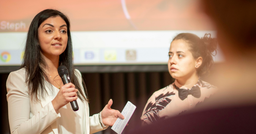 A student stands, microphone in hand, to give a presentation. A projection screen can be seen behind her.