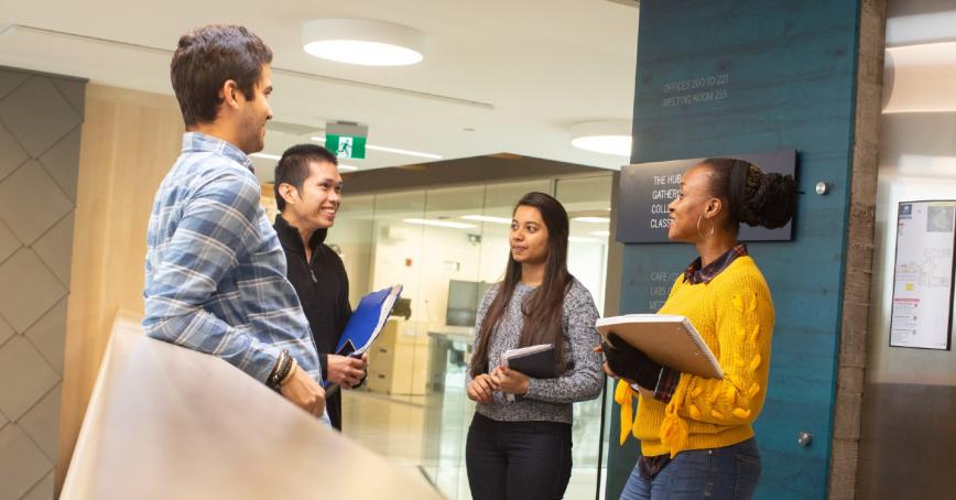 Four students holding notebooks speaking in the hallway 