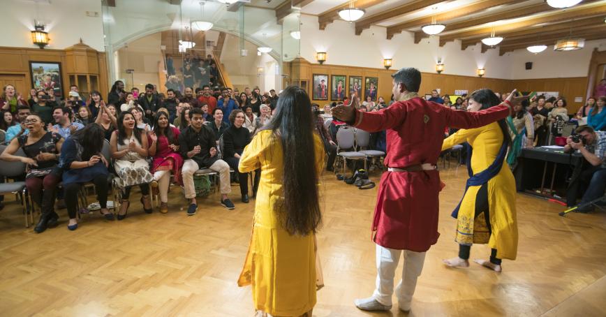Three performers can be seen from behind. An audience watches them.