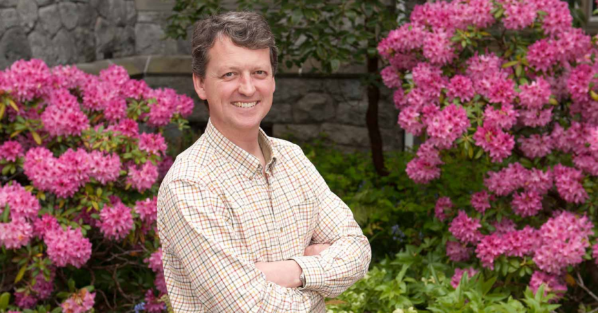Head shot of Geoff Bird standing in front of a stone wall with bushes filled with purple flowers in front