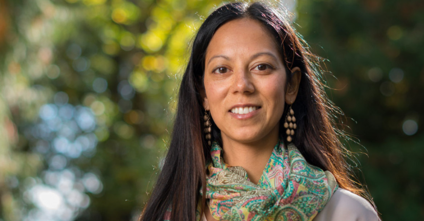 A woman poses for a head shot. She has long brown hair and wears a patterned scarf.