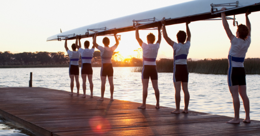 Rowers holding up a boat on a dock with the sun rising in the background