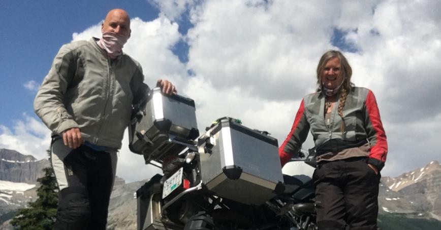 Andreas and Carolann Gneist standing next to a motorcycle with clouds and mountains in the background.