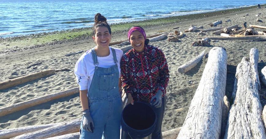 Two BBA students, Emily (left) and Celicia (right) stand on the beach at Esquimalt Lagoon with a bucket for trash