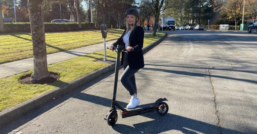 Karly Nygaard-Petersen stands with one foot on an electric scooter on a tree-lined street