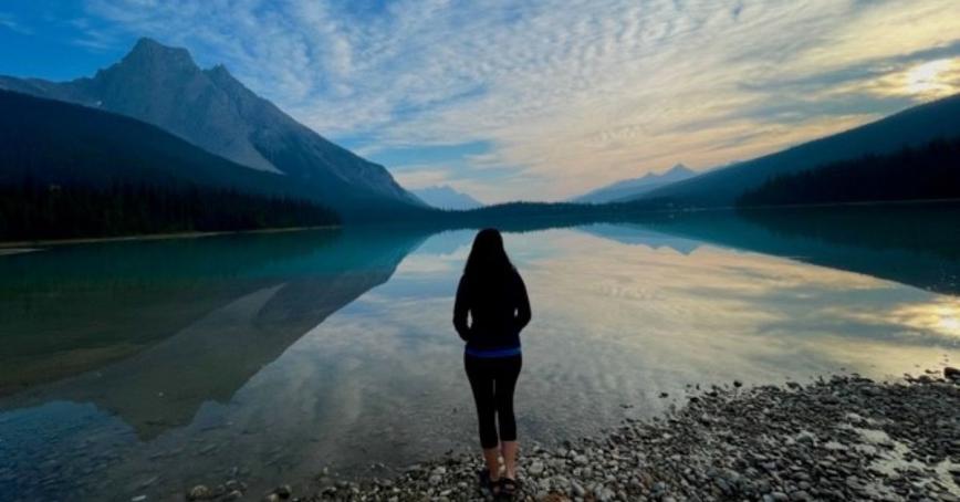 Kate Dunn stands on a rocky beach with a blue lake and mountains behind her