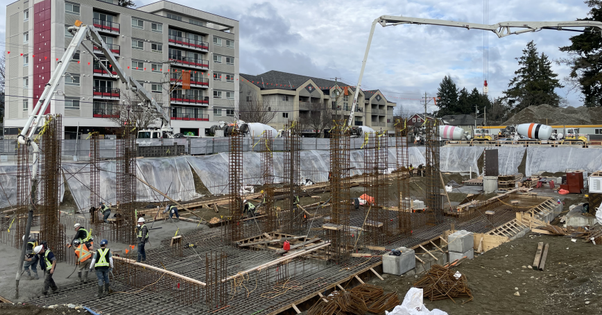 Construction workers aim and pour concrete from an extended concrete pump truck hose into a large excavation site. 