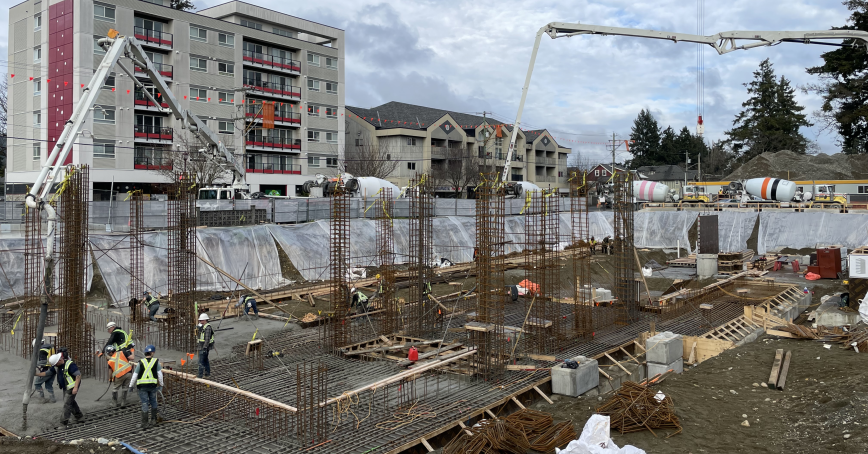 Construction workers aim and pour concrete from an extended concrete pump truck hose into a large excavation site.