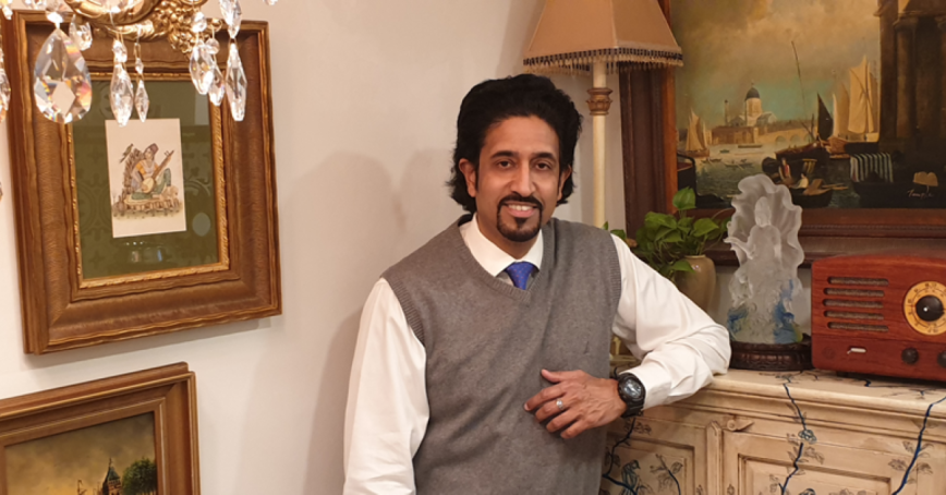 Man wearing sweater vest and dress shirt leans on a piece of furniture. Framed artworks are on the walls.