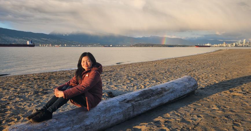 Kim Tholl sits on a log, legs up, on a sandy beach.