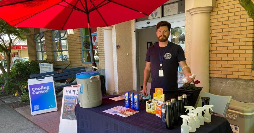 Bearded man standing behind table that has water bottles. There is a sign that says "Cooling Centre" beside the table.