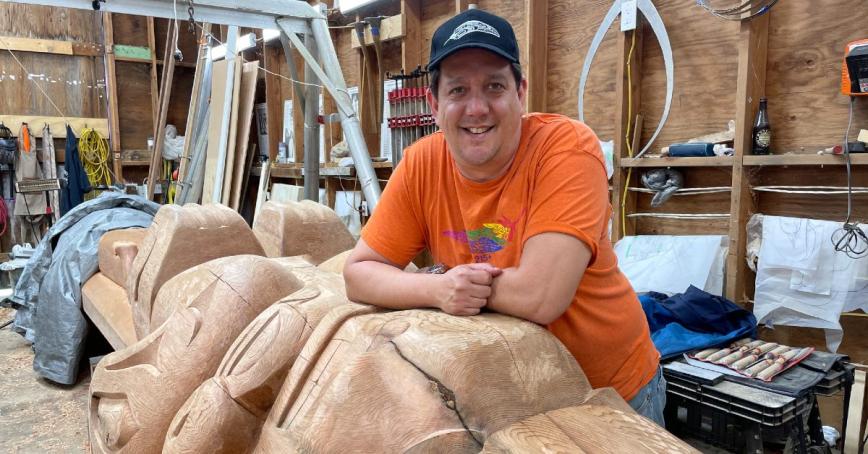 Smiling Indigenous man resting his arms on a partially carved totem pole in a workshop. He is wearing an orange t-shirt and a dark ball cap.