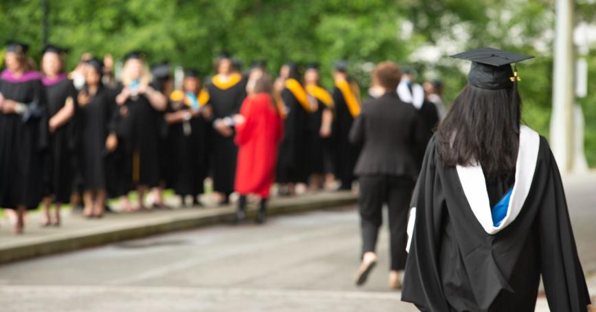 line of grads in robes in the background, with a woman in grad robes in the foreground walking towards them