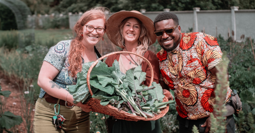 Three people pose for a picture in a garden holding collard greens in a basket.