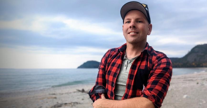 Gary Hayes stands on a beach wearing a flannel shirt and smiling. 