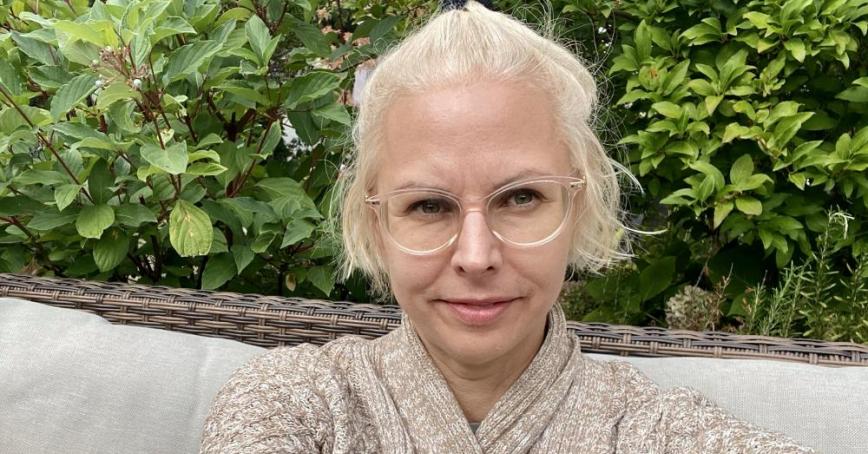 A woman with light blonde hair smiles into the camera in front of a wall of greenery