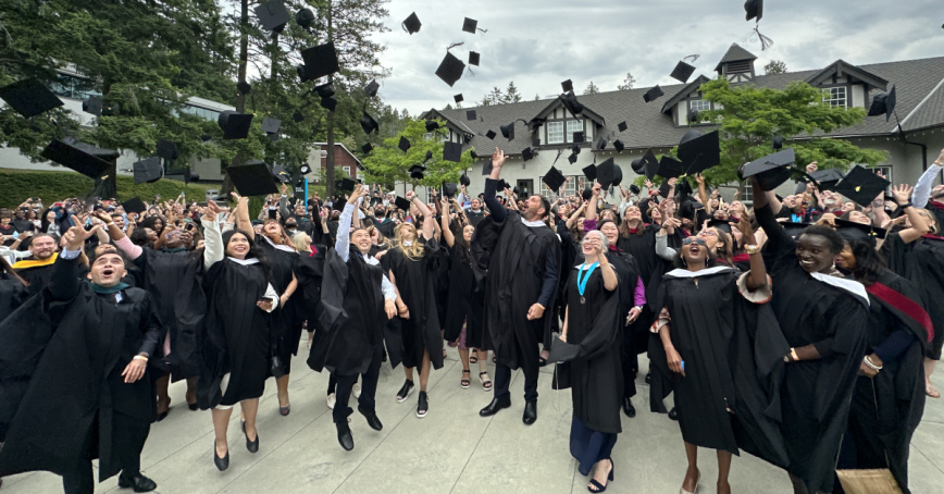 A group of RRU graduates in caps and gowns throw their caps in the air outside of the Sherman Jen Building. 