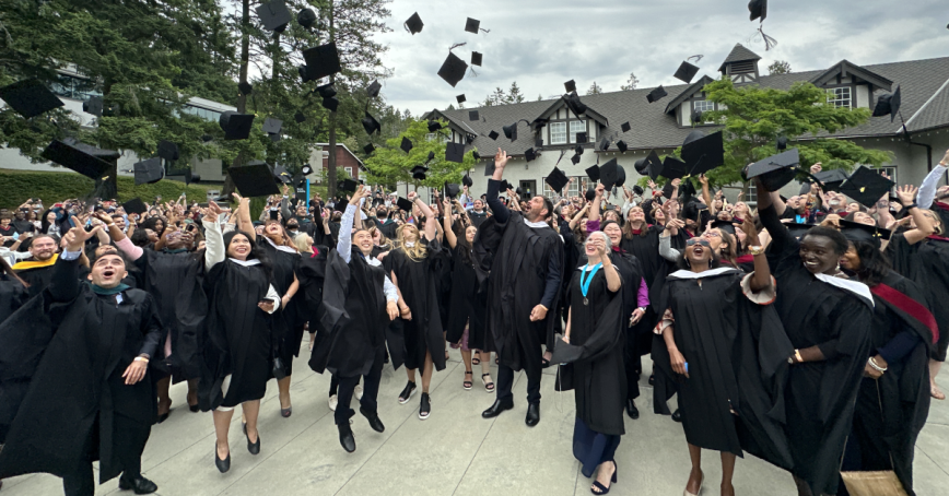 A group of RRU graduates in caps and gowns throw their caps in the air outside of the Sherman Jen Building.