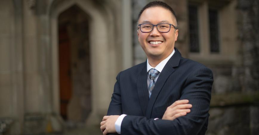 Asian man wearing glasses and a suit smiling at the camera.