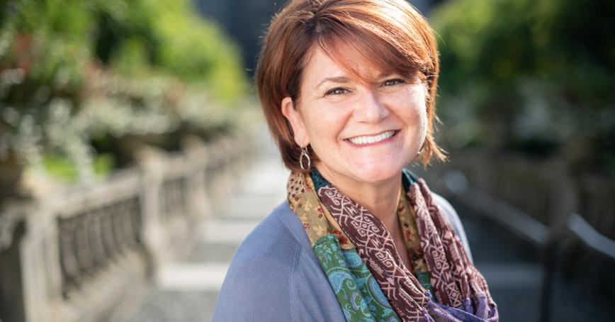Woman with short auburn hair smiling at the camera with trees and stone staircase behind her.