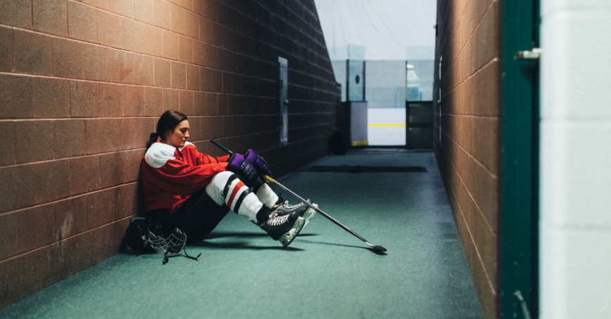young woman in hockey gear sitting on the floor off the ice looking disappointed