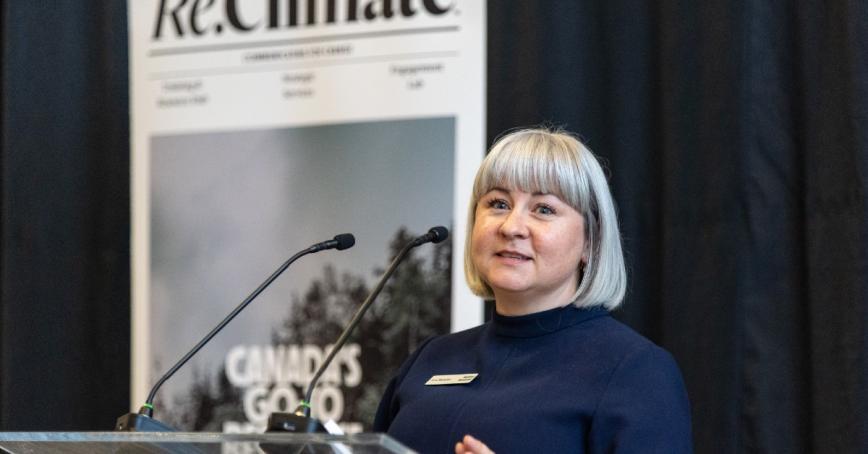Woman with short grey hair and bangs standing at a podium with a sign saying Re,Climate on the wall behind her.