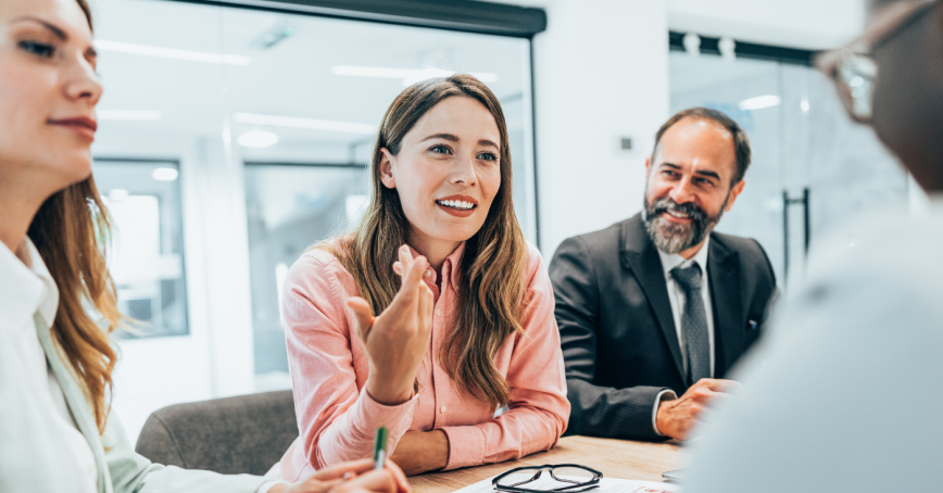 A group of professionals converse around a boardroom table in a brightly lit room. 