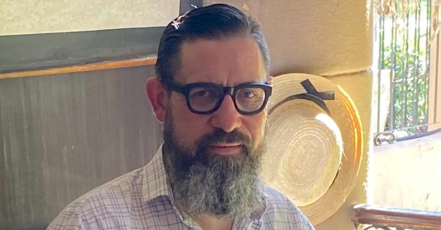 Lew MacDonald is wearing dark rimmed glasses and a white collared shirt. He has a full bear and dark hair.