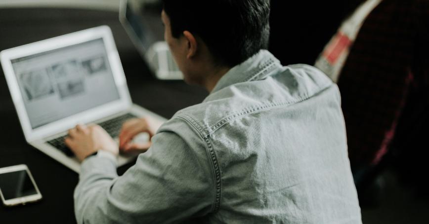 Person sitting at a laptop computer, back to the camera, wear a denim shirt.