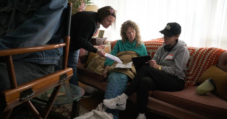 Kim Manky handing papers to a woman sitting on sofa wearing a mermaid costume.