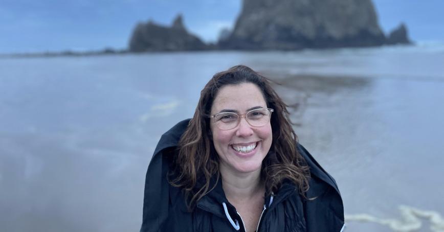 Kyle Epstein smiling and standing on the shore of a body of water with a large rock in the background offshore.